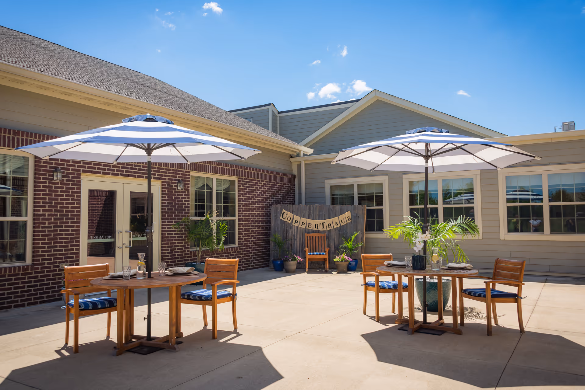 Outdoor patio area at Copper Trace Family-first Senior Living with two round wooden tables, each shaded by a large white and blue striped umbrella. Each table is surrounded by four wooden chairs with blue striped cushions. The patio is paved and bordered by a brick and siding building. A wooden fence with a banner spelling 'COPPER TRACE' is visible in the background, along with potted plants and a wooden chair.
