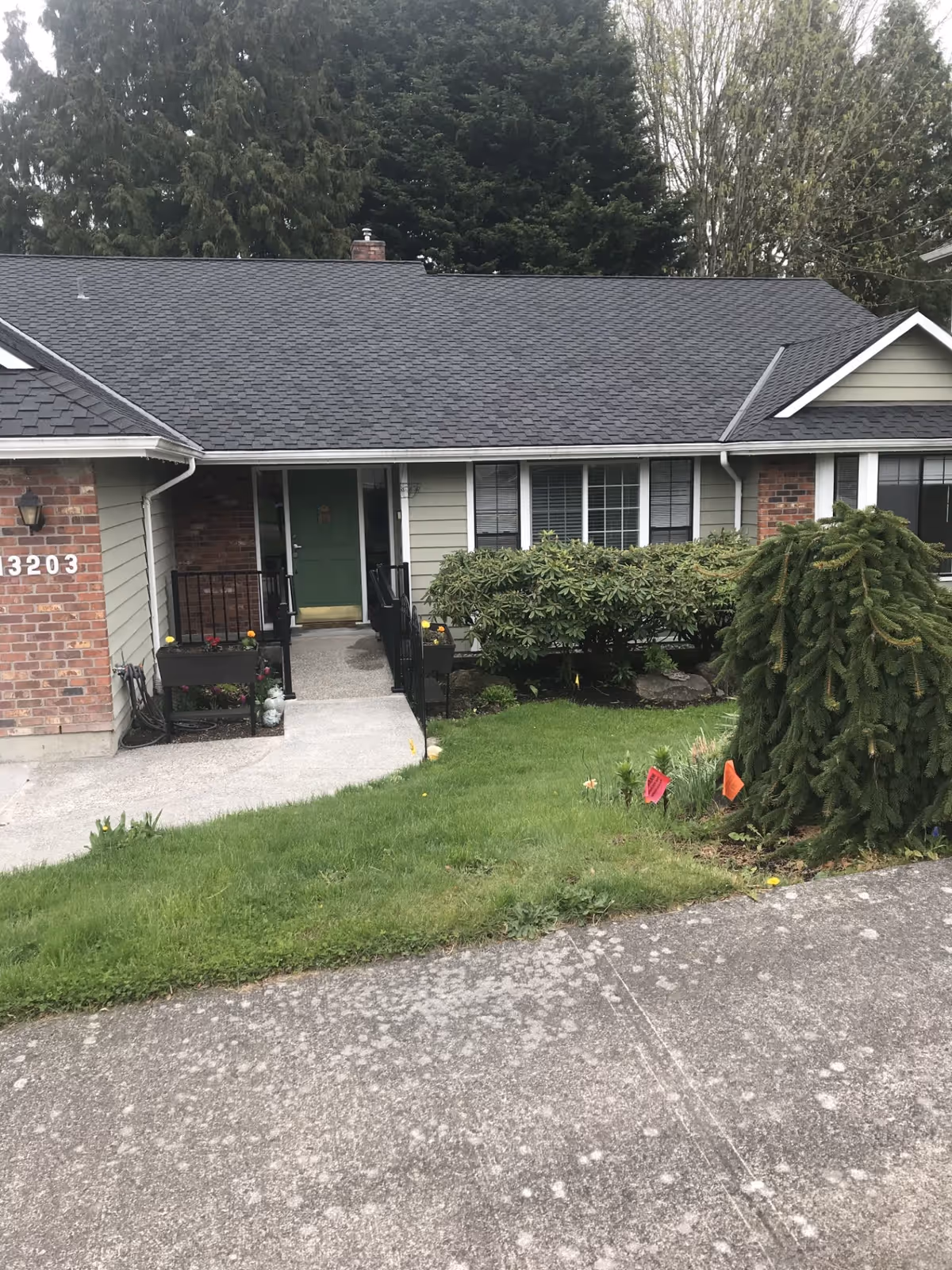 Front exterior view of a single-story house with a green door, brick and siding walls, a small ramp with black railings leading to the entrance, and a well-maintained lawn with bushes and trees.