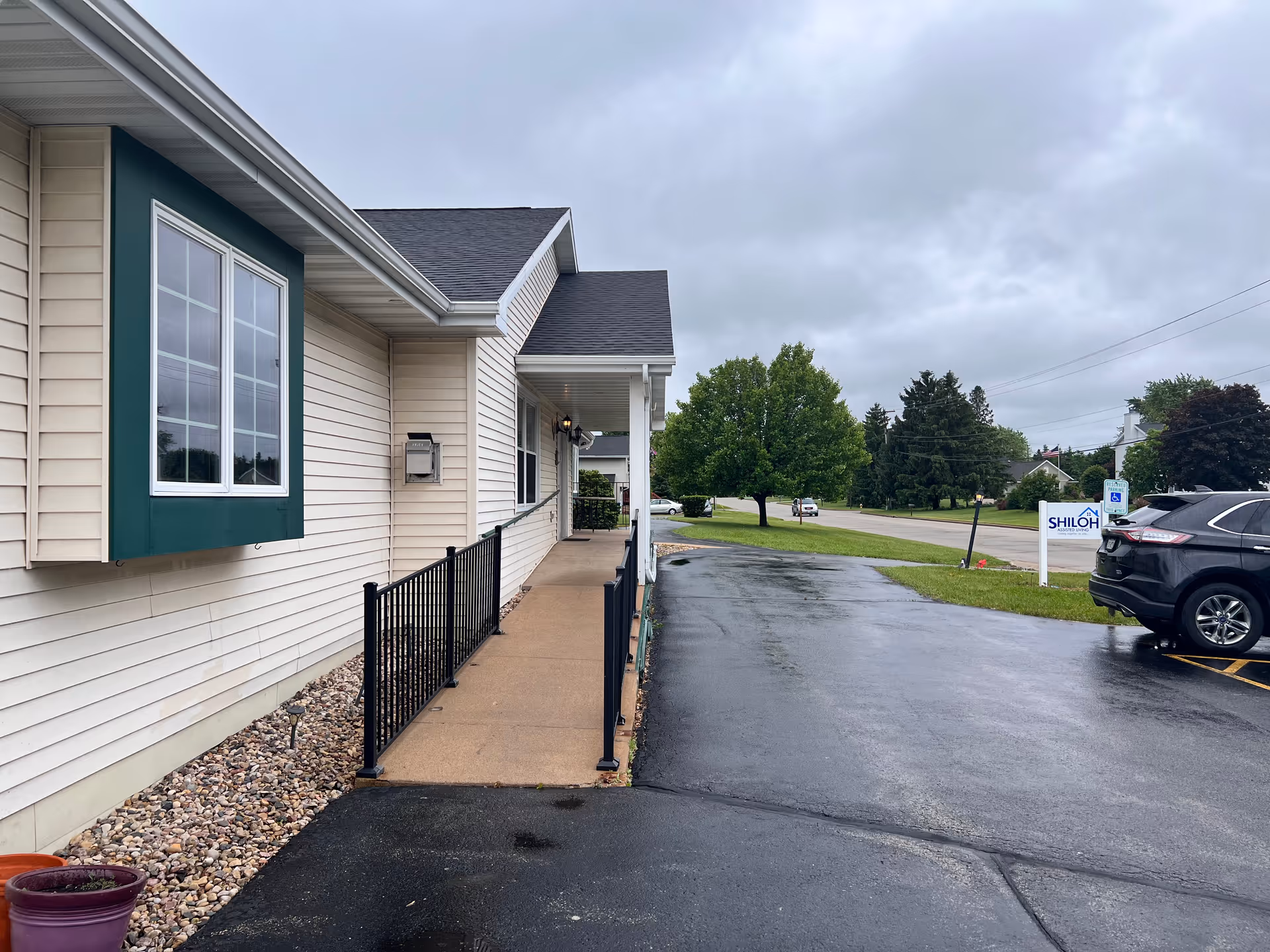 Exterior view of Shiloh Assisted Living facility showing a beige building with green trim around a window, a concrete walkway with black metal railing, a parking lot with a black SUV, and a sign for Shiloh Assisted Living near a grassy area with trees under a cloudy sky.