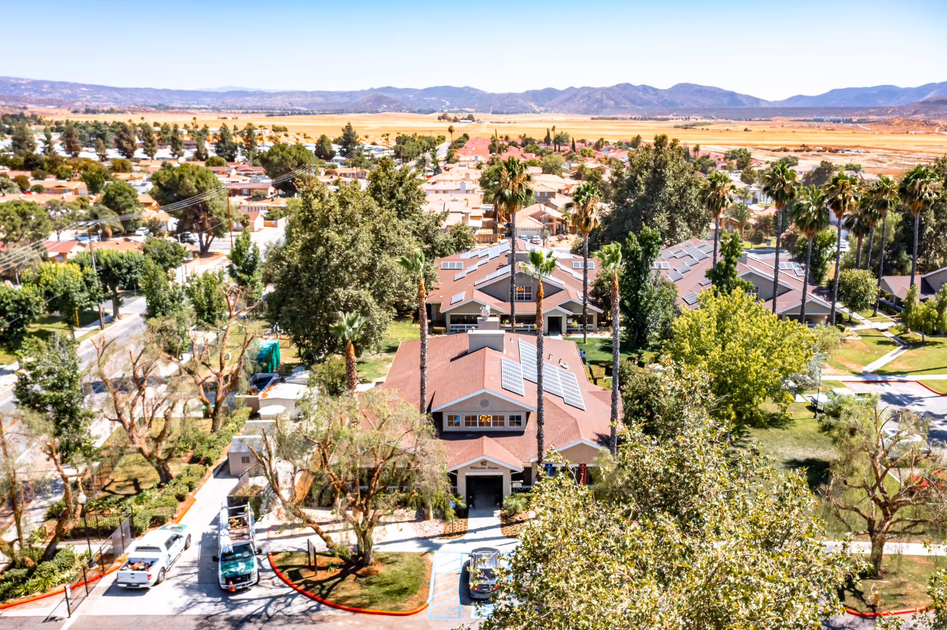 Aerial view of a senior living facility named Cottages at Hemet, showing multiple single-story buildings with brown roofs and solar panels, surrounded by tall palm trees and other greenery. The facility is located in a suburban area with mountains visible in the background under a clear blue sky.