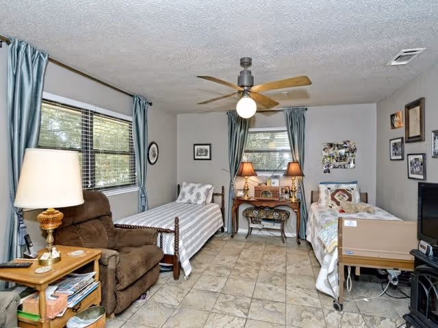 Shared senior bedroom with two twin beds, a recliner, bedside tables, lamps, ceiling fan and windows with blue curtains.