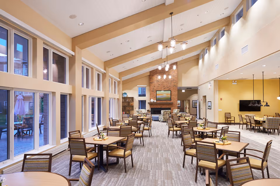 Bright, spacious dining and common room with multiple tables and chairs, large windows, vaulted ceiling, and a brick fireplace.