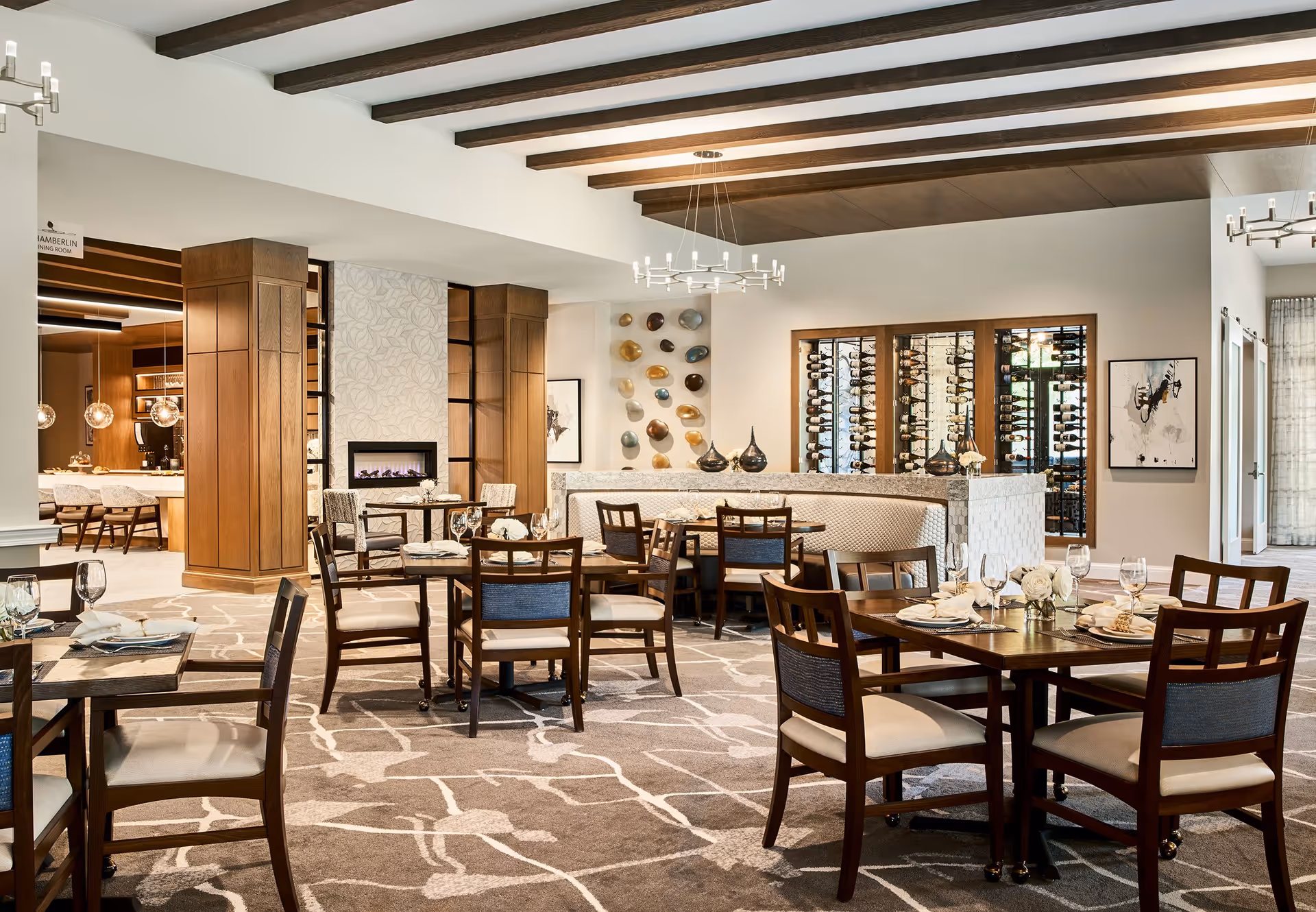 A spacious and elegant dining room with wooden tables and chairs arranged neatly. The room features a patterned carpet, a modern chandelier, a fireplace with a decorative wall, and a wine display cabinet. The ceiling has exposed wooden beams, and the room is well-lit with natural and artificial light.