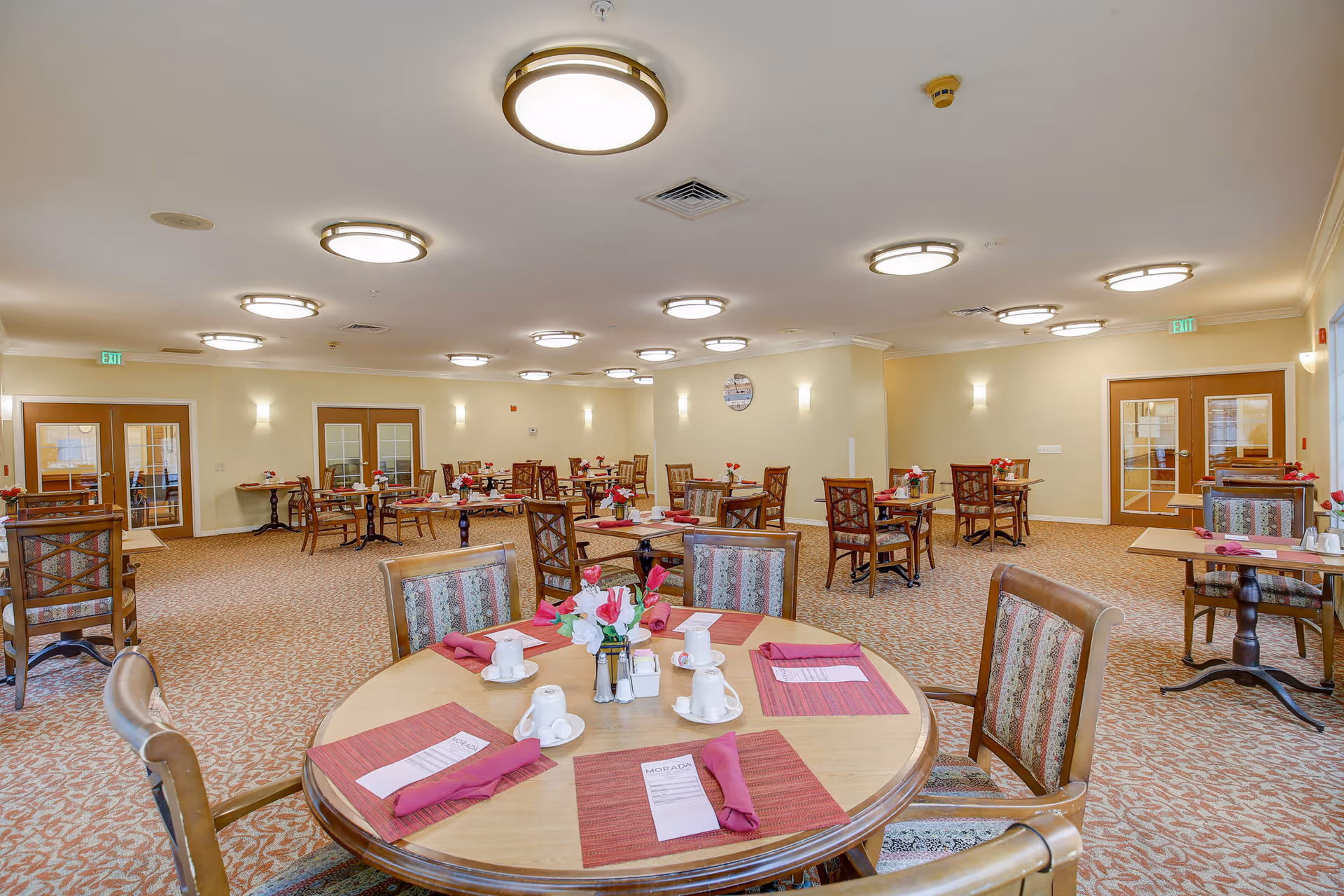 A spacious dining room with multiple round and square wooden tables, each set with red placemats, folded red napkins, white cups, and small flower arrangements. The room has beige walls, carpeted floors with a patterned design, and several ceiling lights. There are double doors on the far walls and exit signs above them.