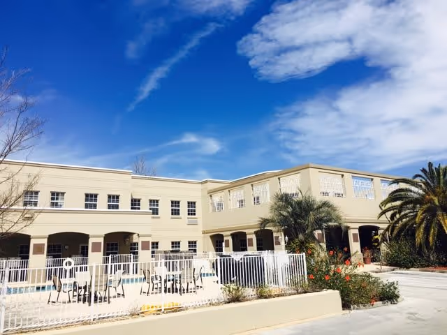 Exterior view of a two-story beige senior living building with a fenced outdoor pool, chairs, and palm trees under a blue sky.