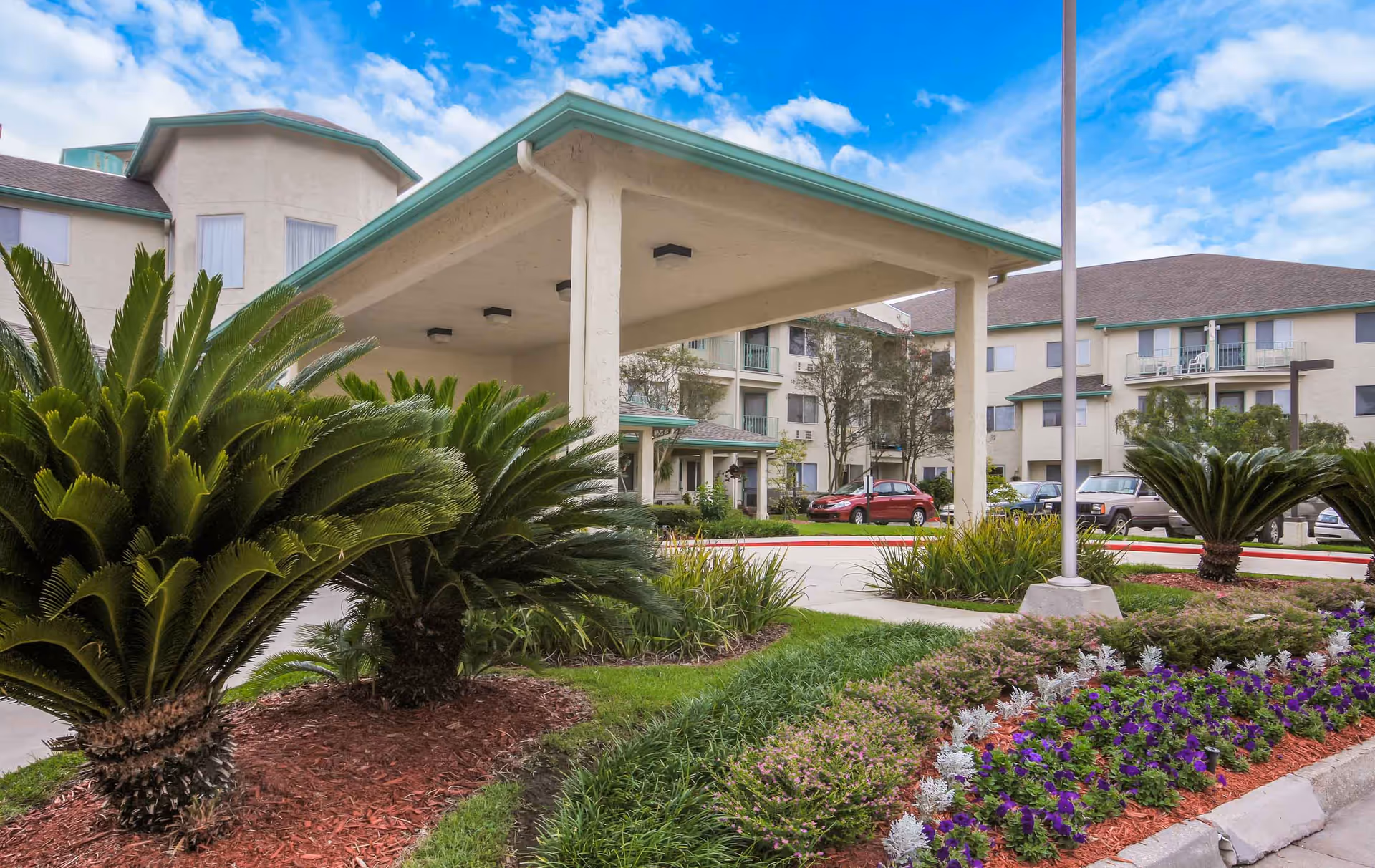 Exterior view of Whealdon Estates by Barclay House showing the entrance canopy, landscaped garden with palm plants and flowers, and a parking area with several cars in front of a multi-story residential building under a partly cloudy blue sky.