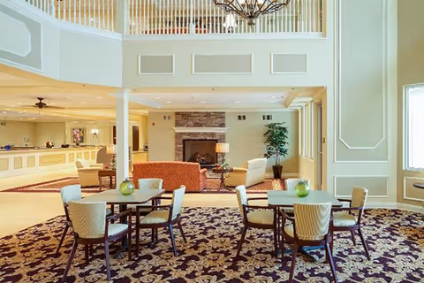 Bright, spacious senior facility common area with small dining tables, upholstered seating, and a fireplace beneath a mezzanine.