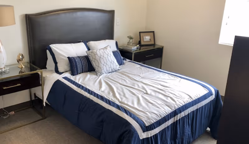 A neatly made bed with a dark upholstered headboard, white and navy blue bedding, and several decorative pillows. There are two bedside tables, one with a lamp and a small decorative item, and the other with books and a framed photo. A window is visible on the right side of the room.