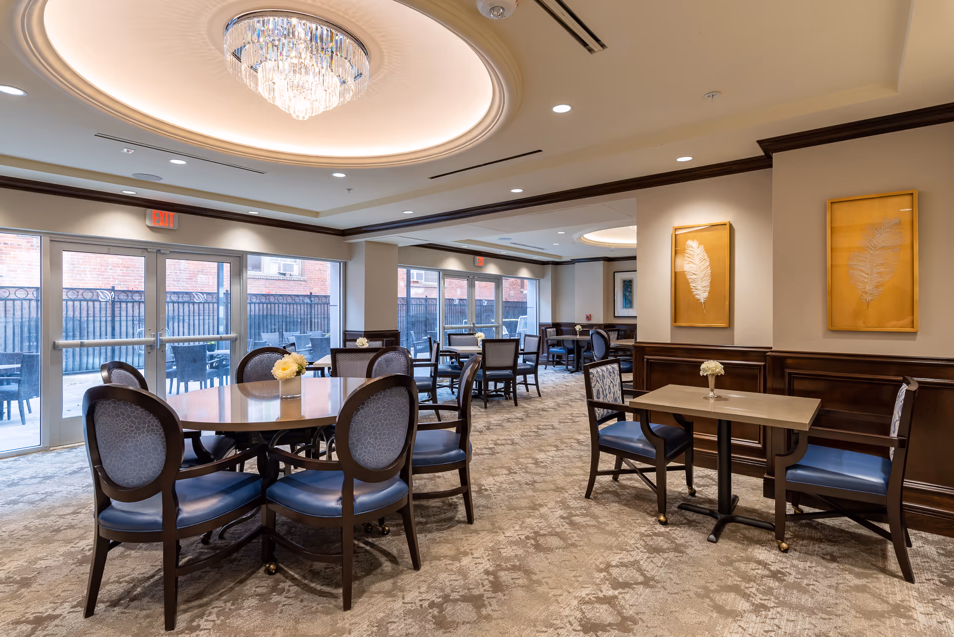 An elegant senior living dining room with round and square tables, blue-upholstered chairs, a central crystal chandelier, and large glass doors leading to an outdoor patio.
