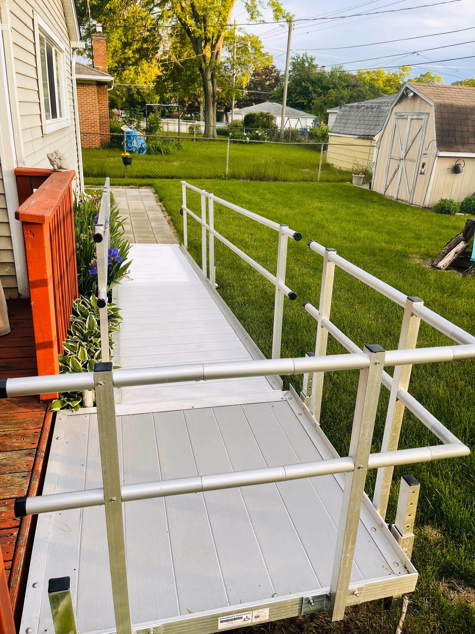 Exterior view of a metal accessibility ramp with handrails leading from a house deck into a grassy backyard with a shed.
