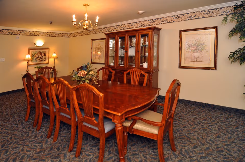 A formal dining room with a large wooden dining table surrounded by eight wooden chairs. The room features a wooden china cabinet filled with dishes and glassware, floral artwork on the walls, carpeted floor, and warm lighting from wall sconces and a chandelier.
