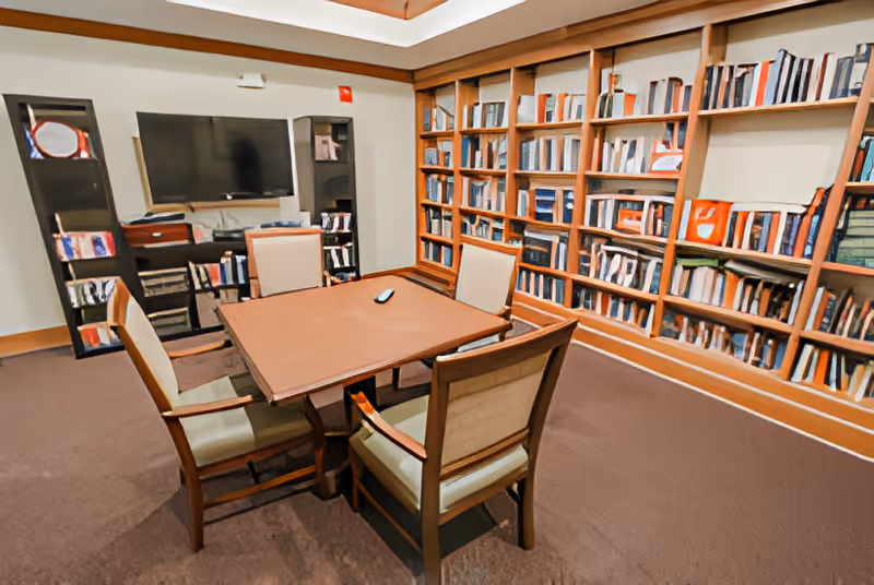 A cozy room with a square wooden table surrounded by four cushioned chairs. The walls are lined with bookshelves filled with books and some decorative items. A large flat-screen TV is mounted on the wall opposite the bookshelves, with additional shelving units below and on the sides holding more books and items. The room has a warm, inviting atmosphere with carpeted flooring and soft lighting.