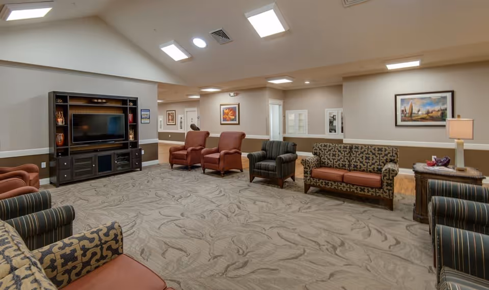 A spacious senior living facility common area with patterned carpet, multiple upholstered chairs and sofas arranged around the room, a large dark wood entertainment center with a flat-screen TV, framed artwork on the walls, and ceiling lights providing bright illumination.