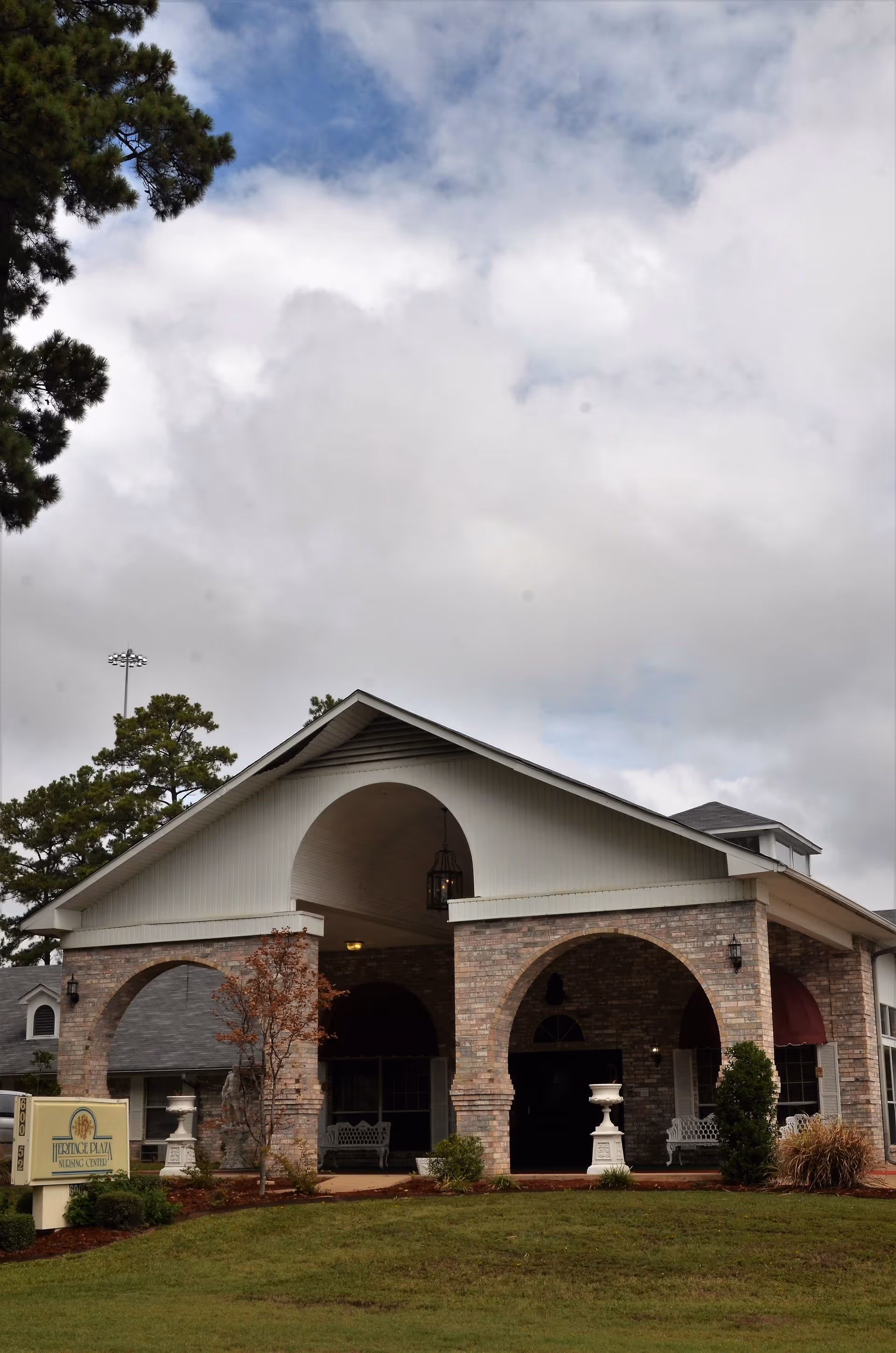 Exterior view of Heritage Plaza Nursing Center showing a brick building with large arched entryways, a covered porch area with benches, decorative urns, and a sign with the facility's name on a grassy lawn under a cloudy sky.