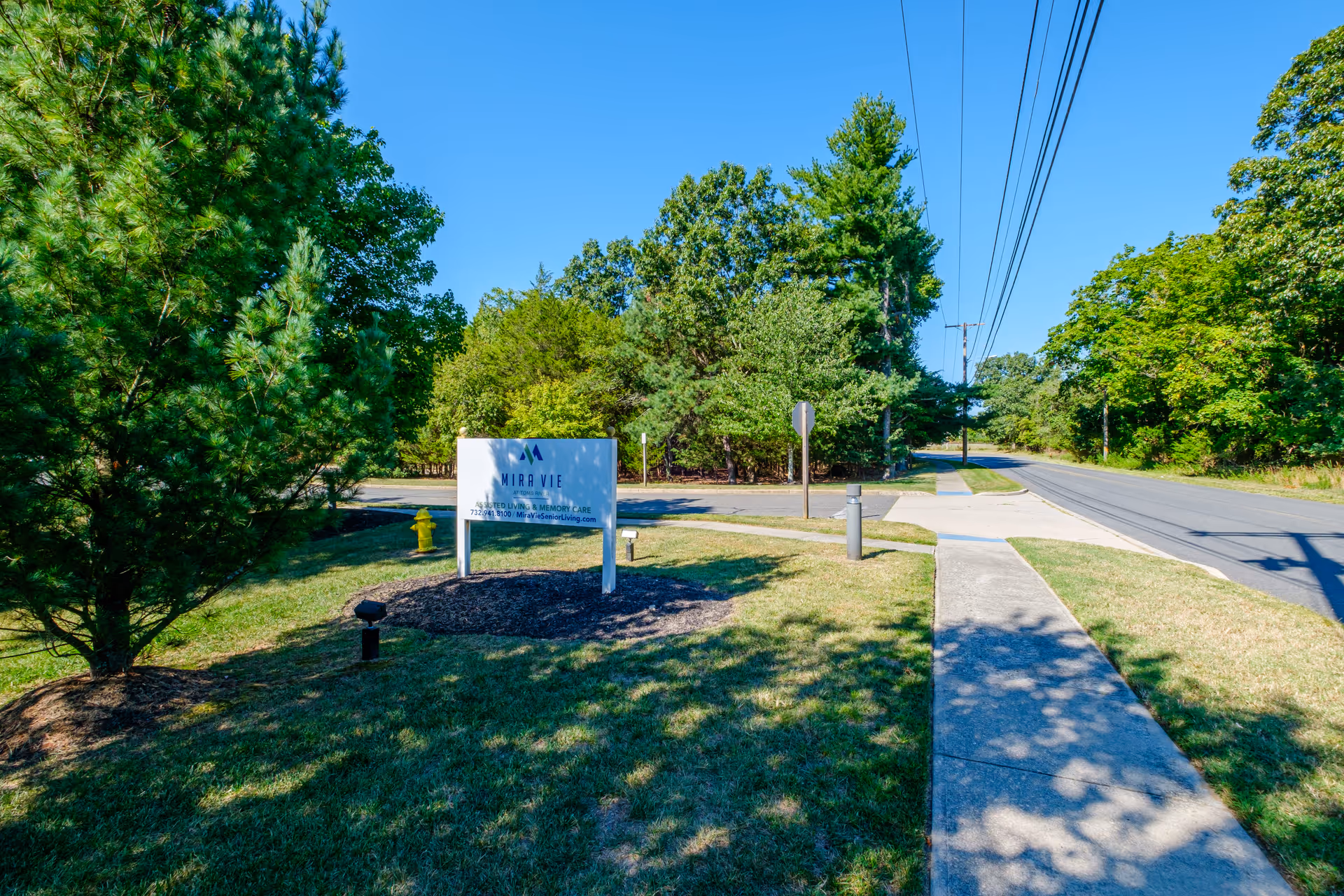 Outdoor view of a sidewalk leading to a street with a sign for Mira Vie at Toms River assisted living and memory care facility surrounded by green trees and grass under a clear blue sky.