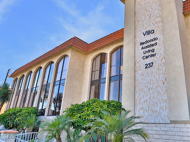 Front exterior of the Villa Redondo Assisted Living Center with tall arched windows, landscaping, and building signage.