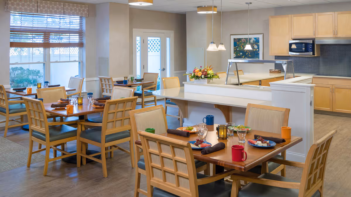 A bright dining room in a senior living facility with wooden tables and chairs. Tables are set with plates of food, glasses, mugs, and napkins. The room has large windows with blinds, a serving counter with a flower arrangement, and a kitchen area with wooden cabinets, a microwave, and a tiled backsplash.