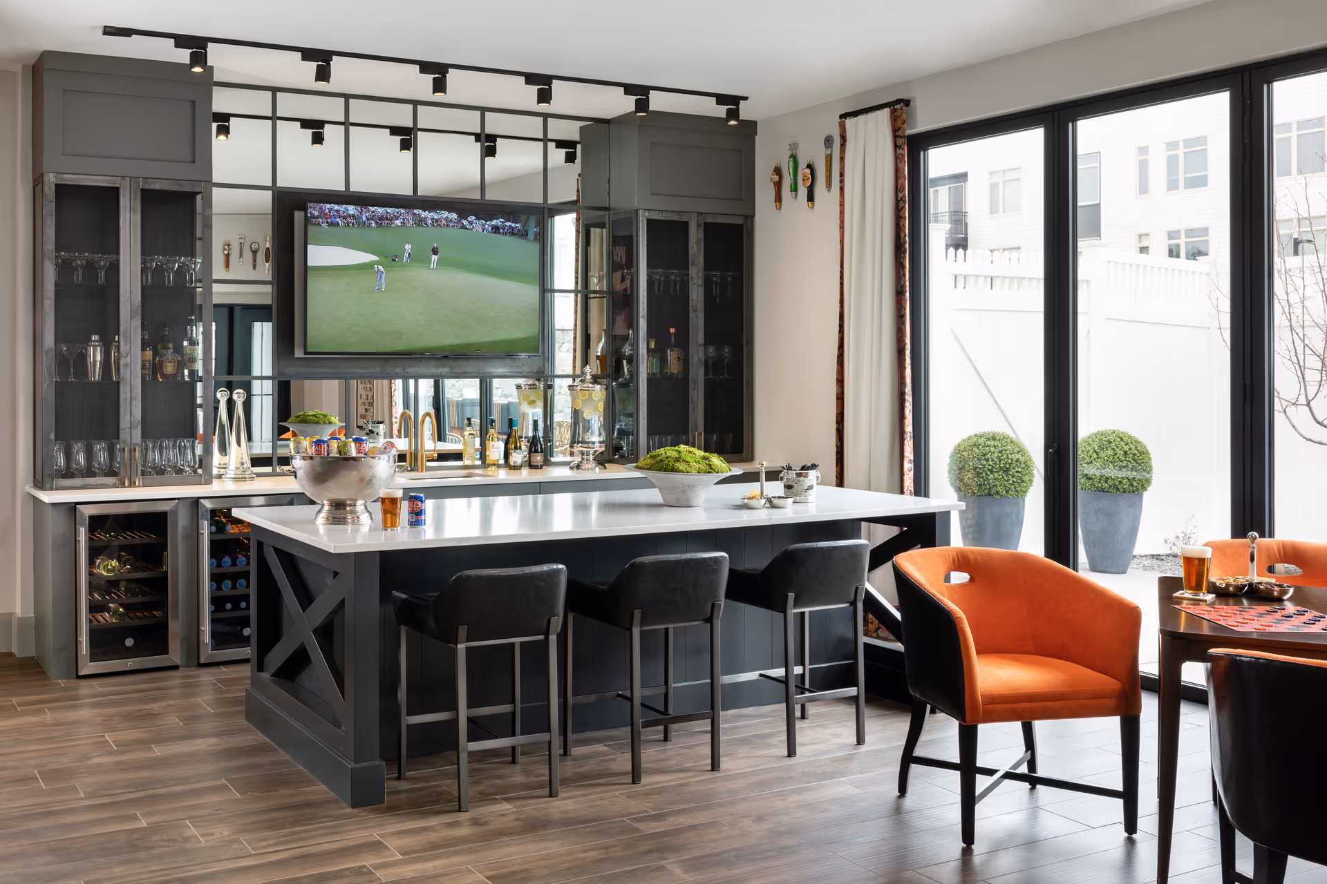 Bright communal lounge and bar area featuring a central island with three barstools, a wall-mounted TV, and seating by floor-to-ceiling glass doors.
