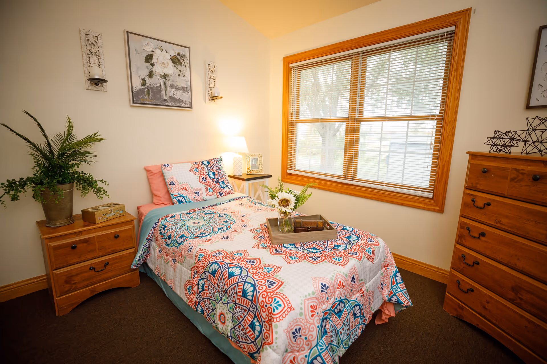 A bright single bedroom with a colorful patterned bedspread, wooden nightstand and dresser, and a window with blinds.