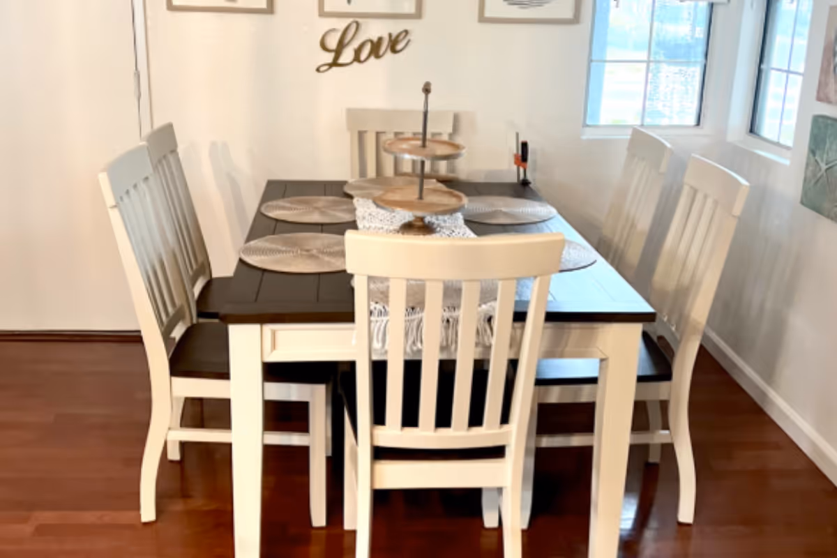 A dining room with a rectangular wooden table and six white chairs. The table has a dark top and white legs, with a three-tiered wooden tray centerpiece and round placemats. The room has wooden flooring, white walls, and two windows letting in natural light. The word 'Love' is displayed on the wall above the table.