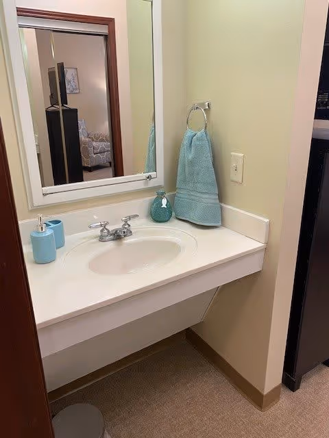 A small bathroom sink area with a white countertop and built-in sink. There is a silver faucet, a blue soap dispenser, a blue cup, and a blue hand towel hanging on a silver ring attached to the wall. A mirror is mounted above the sink, reflecting part of a room with a patterned armchair and a TV. The walls are painted light yellow, and the floor is carpeted.