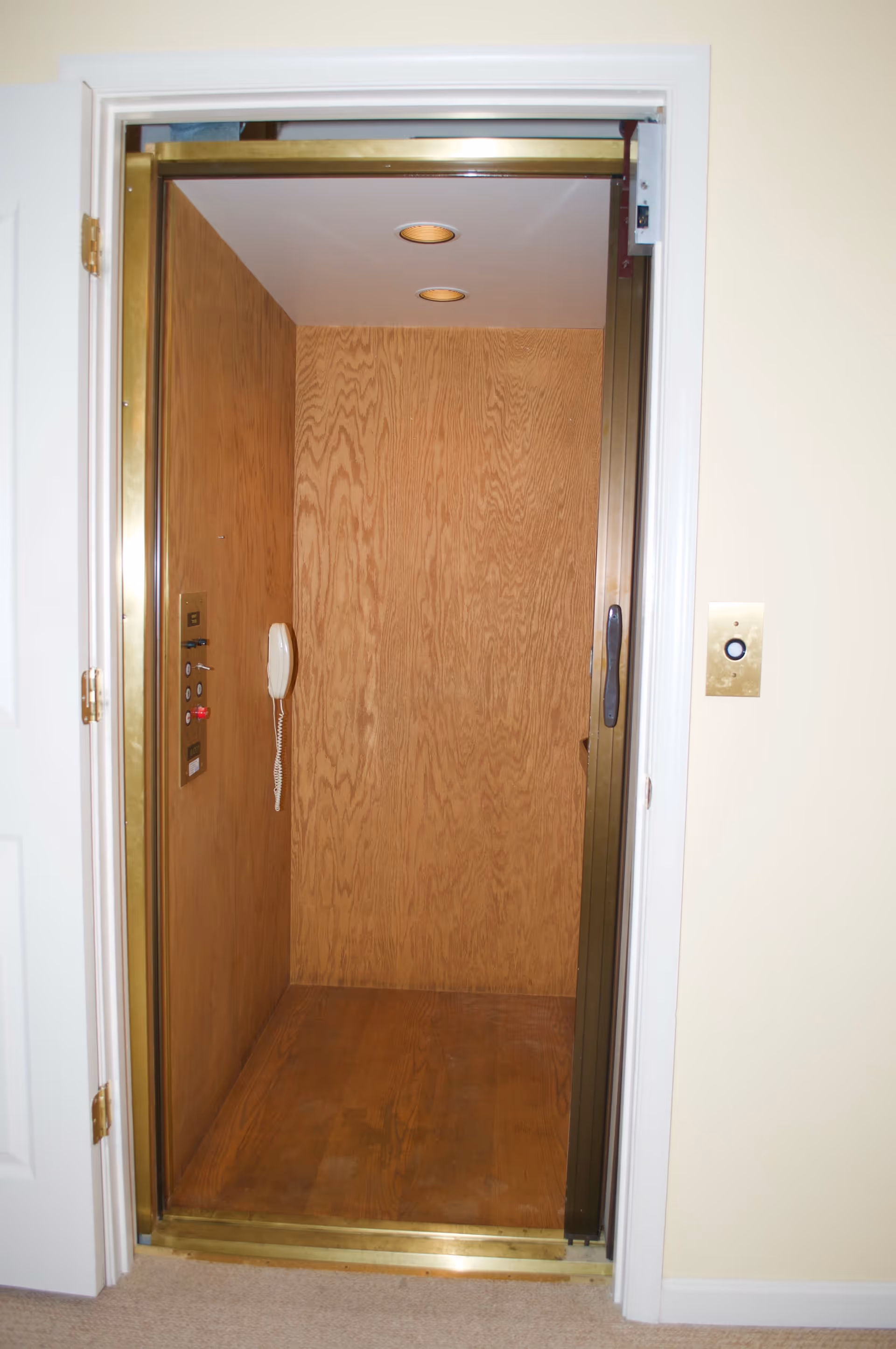 Open residential elevator with wood-paneled interior walls and floor, brass trim, two ceiling lights, and a wall-mounted telephone handset inside. The elevator door is open, showing a beige carpeted floor outside.