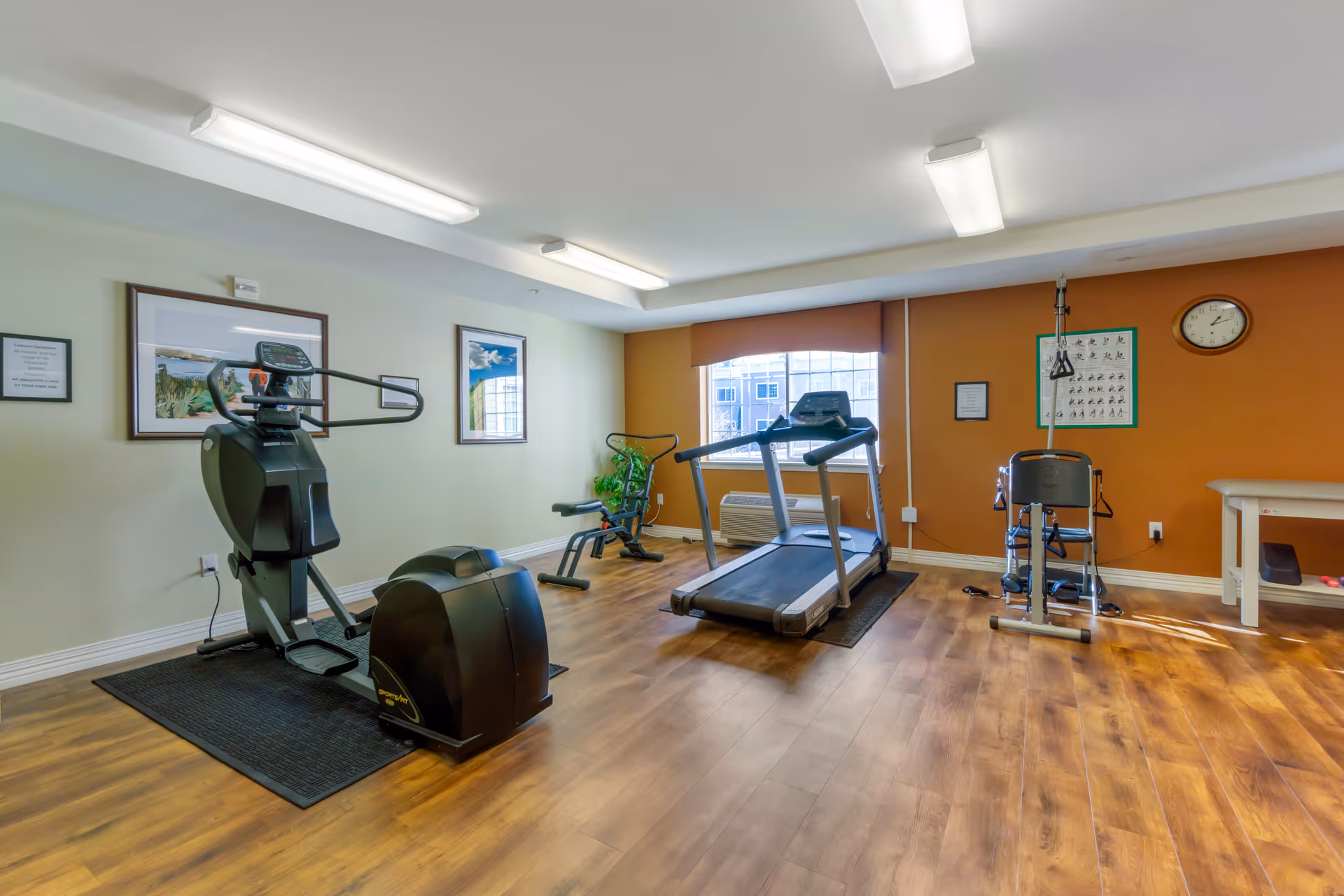 A small fitness room with exercise equipment including a recumbent bike, treadmill, and a multi-functional exercise machine. The room has wood flooring, white and orange walls, framed pictures, a clock, and a window with a view of buildings outside.