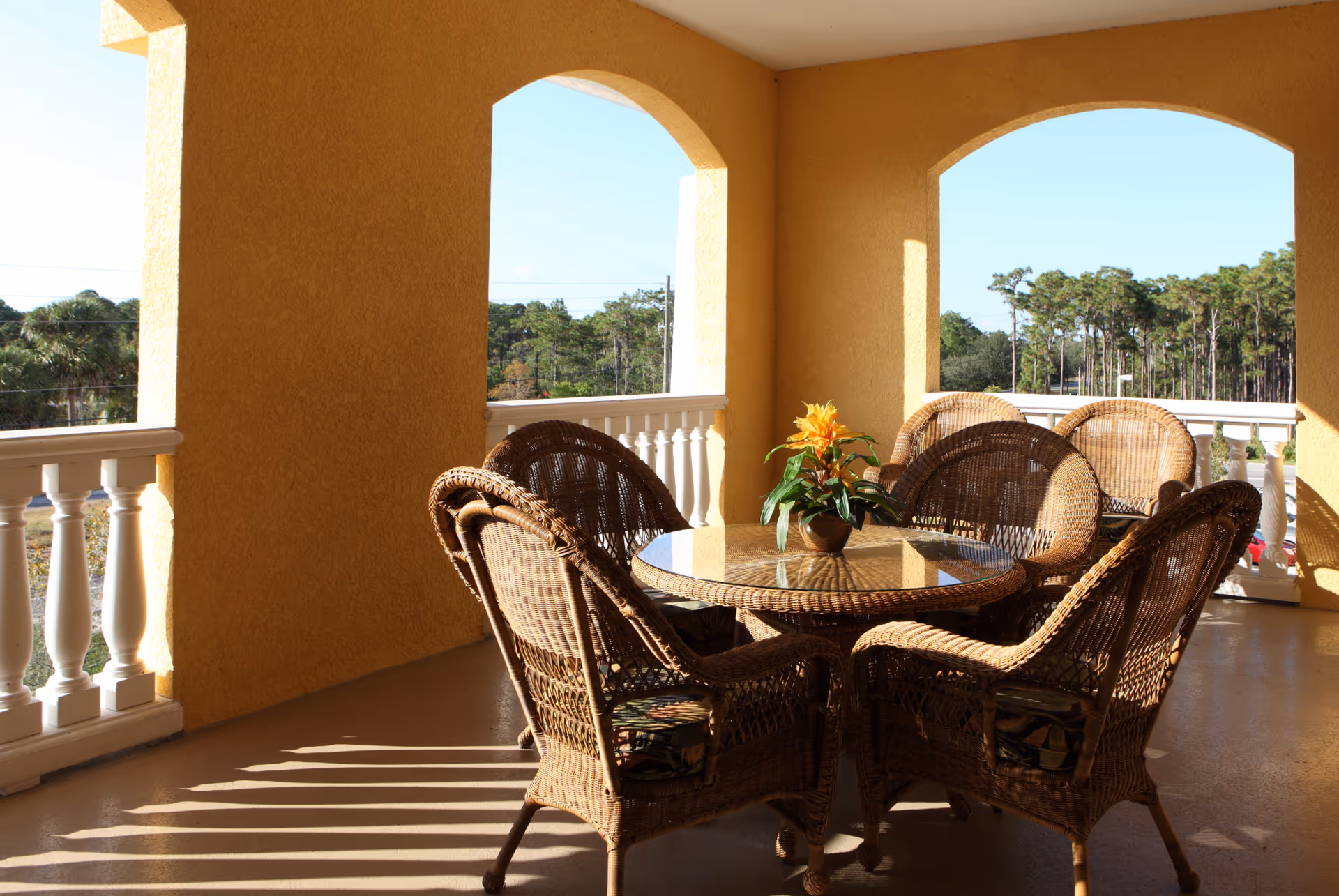 Outdoor covered patio area with yellow textured walls and white balustrades. A round glass-top wicker table is surrounded by six wicker chairs with cushions. A potted plant with yellow flowers is placed in the center of the table. Trees and a clear blue sky are visible in the background.