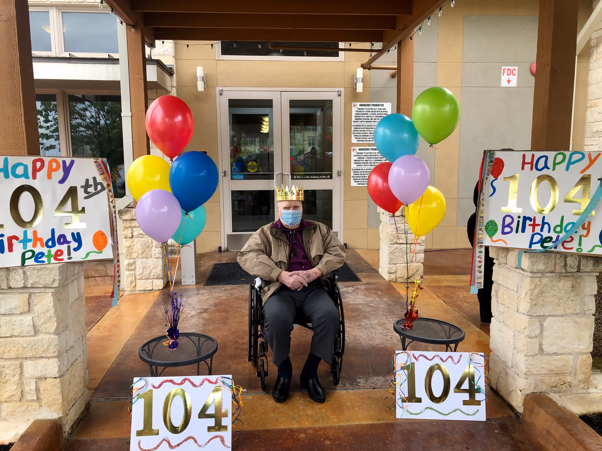An elderly man wearing a crown and a face mask sits in a wheelchair outside the entrance of a building. He is surrounded by colorful balloons and signs celebrating his 104th birthday, with messages saying 'Happy 104th Birthday Pete!'