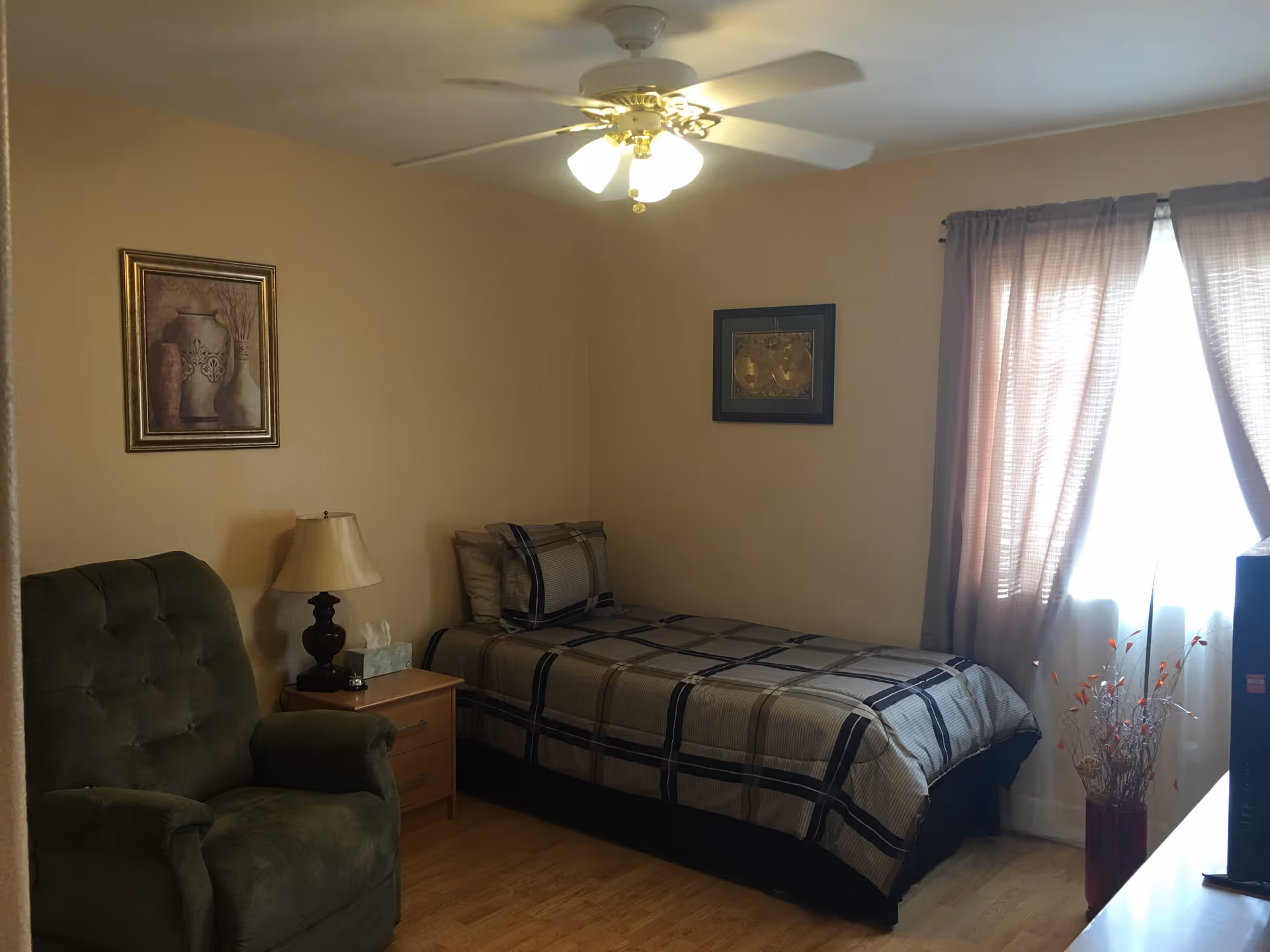 A small furnished bedroom with a single bed, green recliner, nightstand and lamp, ceiling fan, and a curtained window.