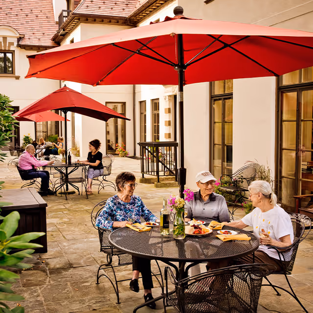 Three elderly people sitting at a round metal table with a red umbrella overhead, enjoying a meal and conversation on a stone patio outside a building. In the background, two other people are seated at another table with a similar red umbrella. The setting is bright and inviting with flowers on the tables and large windows on the building.