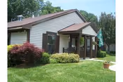 Single-story building with beige siding and brown trim surrounded by green grass and shrubs, with a small porch and a green flag near the entrance under a clear sky.