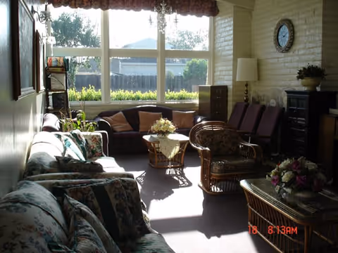 A cozy living room with multiple floral and patterned sofas and chairs arranged around a central coffee table with a flower arrangement. Large windows let in natural light and offer a view of greenery outside. The room features a brick wall with a clock, a floor lamp, and additional seating along the wall.