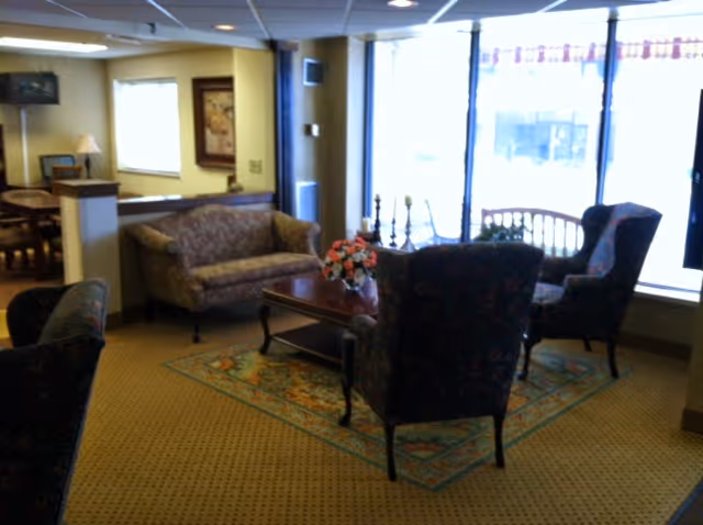 A cozy senior living facility lounge area with patterned armchairs and a sofa arranged around a wooden coffee table with a flower arrangement. The room has large windows letting in natural light, a carpeted floor, and warm yellow walls with framed artwork.