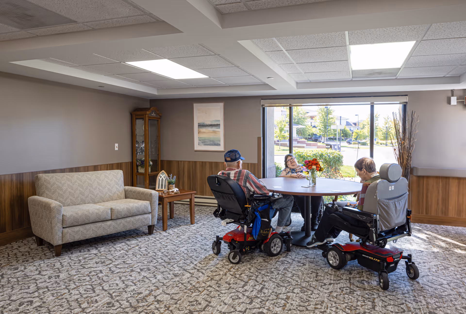 A bright room with a large window showing an outdoor view. Inside, three elderly people are seated around a round table with a vase of flowers. Two of them are in motorized wheelchairs, and one is seated on a regular chair. The room has a patterned carpet, a beige couch, a wooden side table, a glass cabinet, and a framed painting on the wall.