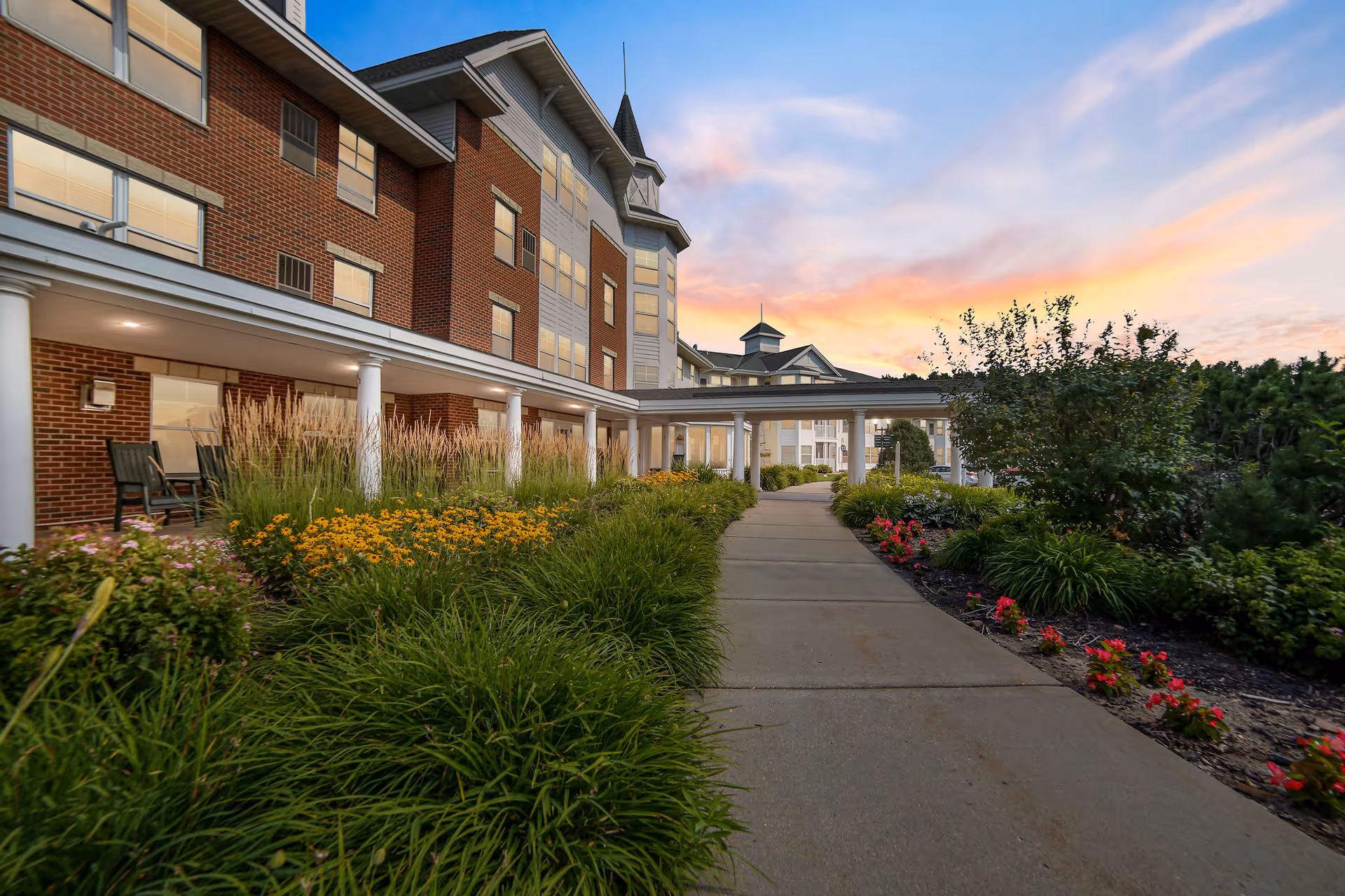 A paved walkway leading alongside a multi-story brick and siding building with white columns and a covered entrance. The walkway is bordered by lush green plants and colorful flowers, with a sunset sky in the background.