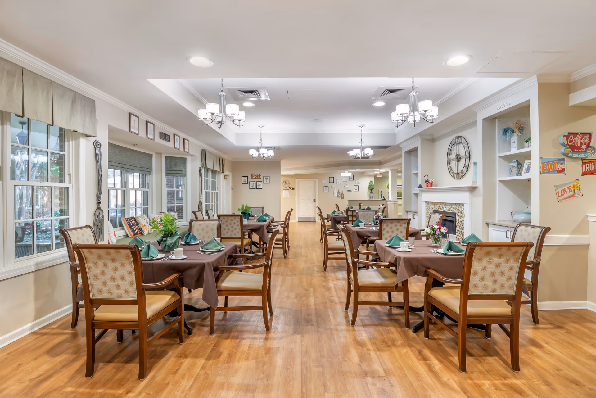 A bright and inviting dining room in a senior living facility with several tables covered in brown tablecloths, each set with green napkins, cups, and plates. Wooden chairs with cushioned seats surround the tables. Large windows on the left side let in natural light, and the walls are decorated with framed pictures and signs with words like 'Eat' and 'Coffee'. The room features wooden flooring, chandeliers, and a fireplace with a large clock above it.