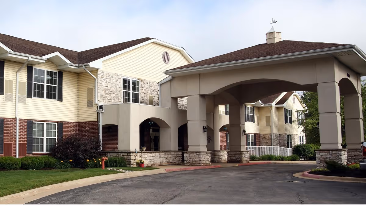 Exterior front entrance of a senior living building with a covered porte-cochere, driveway, and landscaped grounds.