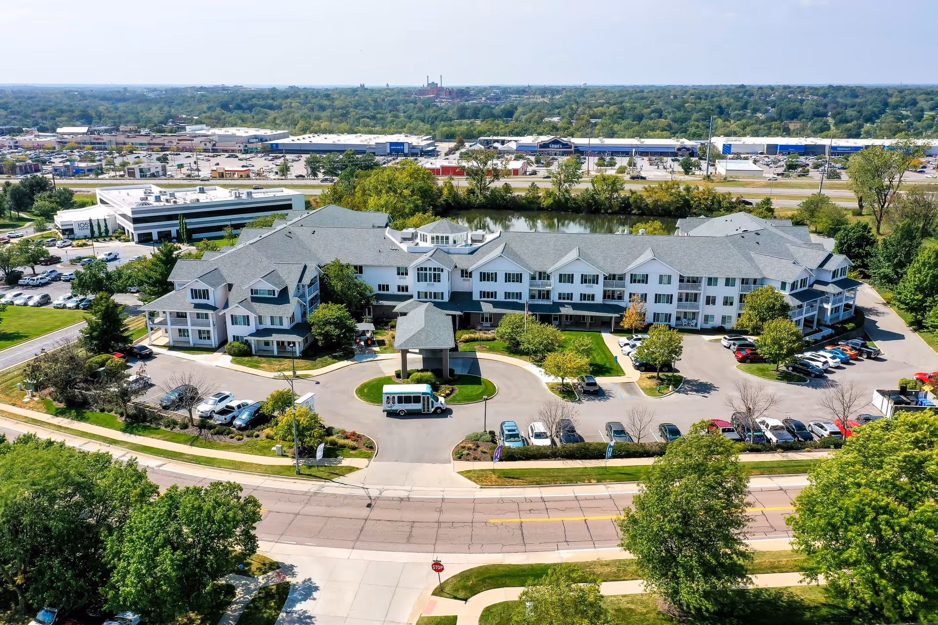 Aerial view of a large senior living facility building with multiple floors, surrounded by parking lots, trees, and greenery. A small shuttle bus is parked in front of the main entrance with a covered drop-off area. In the background, there is a pond and commercial buildings along a highway.