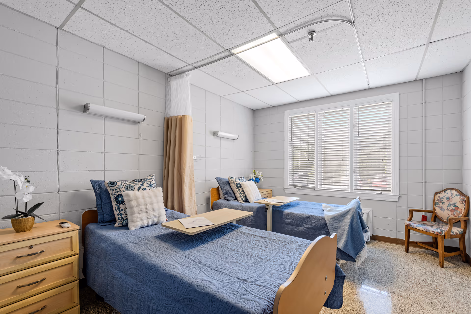 A bright room with two single beds covered in blue bedspreads and decorative pillows. Each bed has an overbed table with papers on it. There is a wooden nightstand with a potted orchid and a remote control between the beds. A floral upholstered wooden chair is placed near a window with white blinds. The walls are light gray with a tiled pattern, and the ceiling has fluorescent lighting and a curtain track with a beige privacy curtain partially drawn.