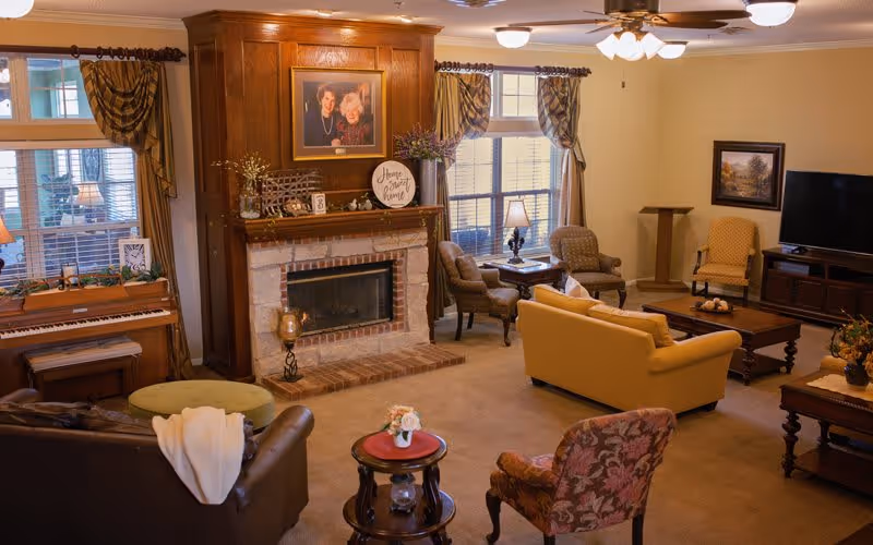 A cozy senior living community common area with a brick fireplace surrounded by wooden paneling, a piano near the window, multiple armchairs and sofas arranged around coffee tables, warm lighting, and decorative curtains on the windows.
