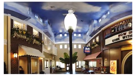 Indoor common area designed to resemble a small town street with storefronts, including a cafe and a theater marquee. The ceiling is painted to look like a blue sky with clouds, and there is a large streetlamp in the center. Seating areas and plants are visible along the sides.