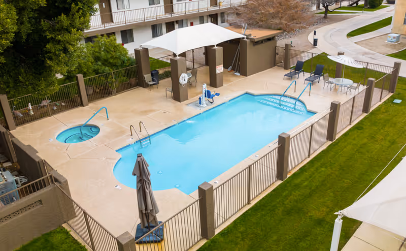 A fenced outdoor swimming pool area at Desert Marigold Senior Living of Tempe, featuring a rectangular pool with steps, a small round hot tub, several lounge chairs, a table with chairs, and shaded seating areas. The pool area is surrounded by a concrete deck and green grass.