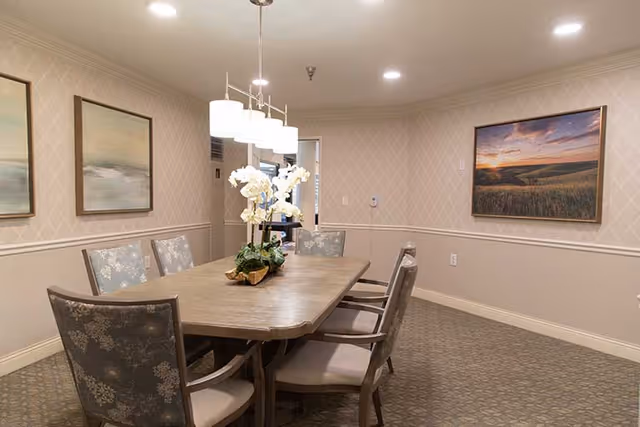 A dining room with a rectangular wooden table surrounded by six cushioned chairs with floral upholstery. A centerpiece with white flowers is placed on the table. The walls are decorated with patterned wallpaper and three framed landscape paintings. A modern light fixture with five lamps hangs above the table.
