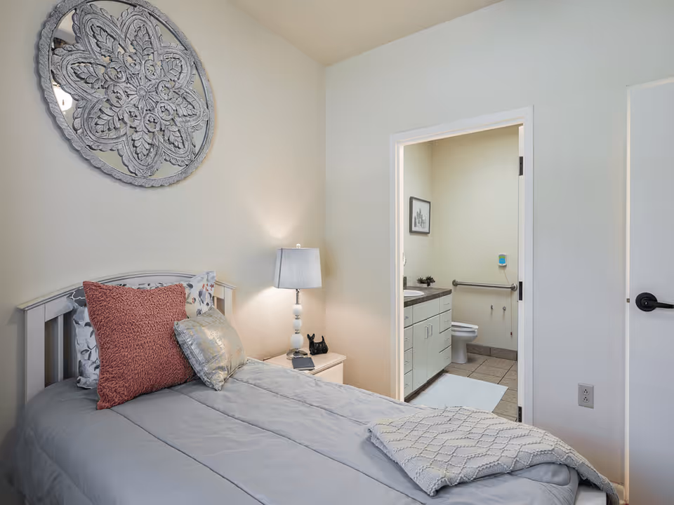 A small bedroom with a single bed made with gray bedding and decorative pillows. A bedside table with a lamp and small decor is next to the bed. A decorative round mirror hangs on the wall above the bed. An open door reveals an adjacent bathroom with a toilet, sink, and grab bar.