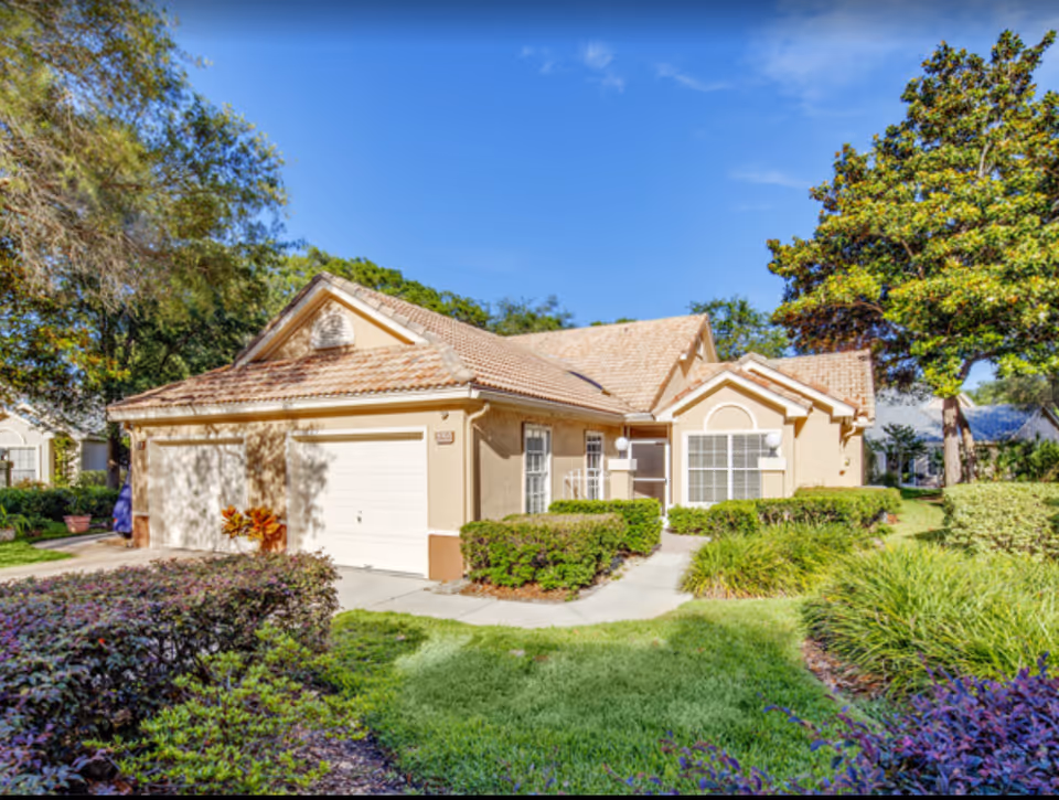 Exterior view of a single-story residential building with a tiled roof, beige walls, and a two-car garage. The building is surrounded by well-maintained green lawns, bushes, and trees under a clear blue sky.
