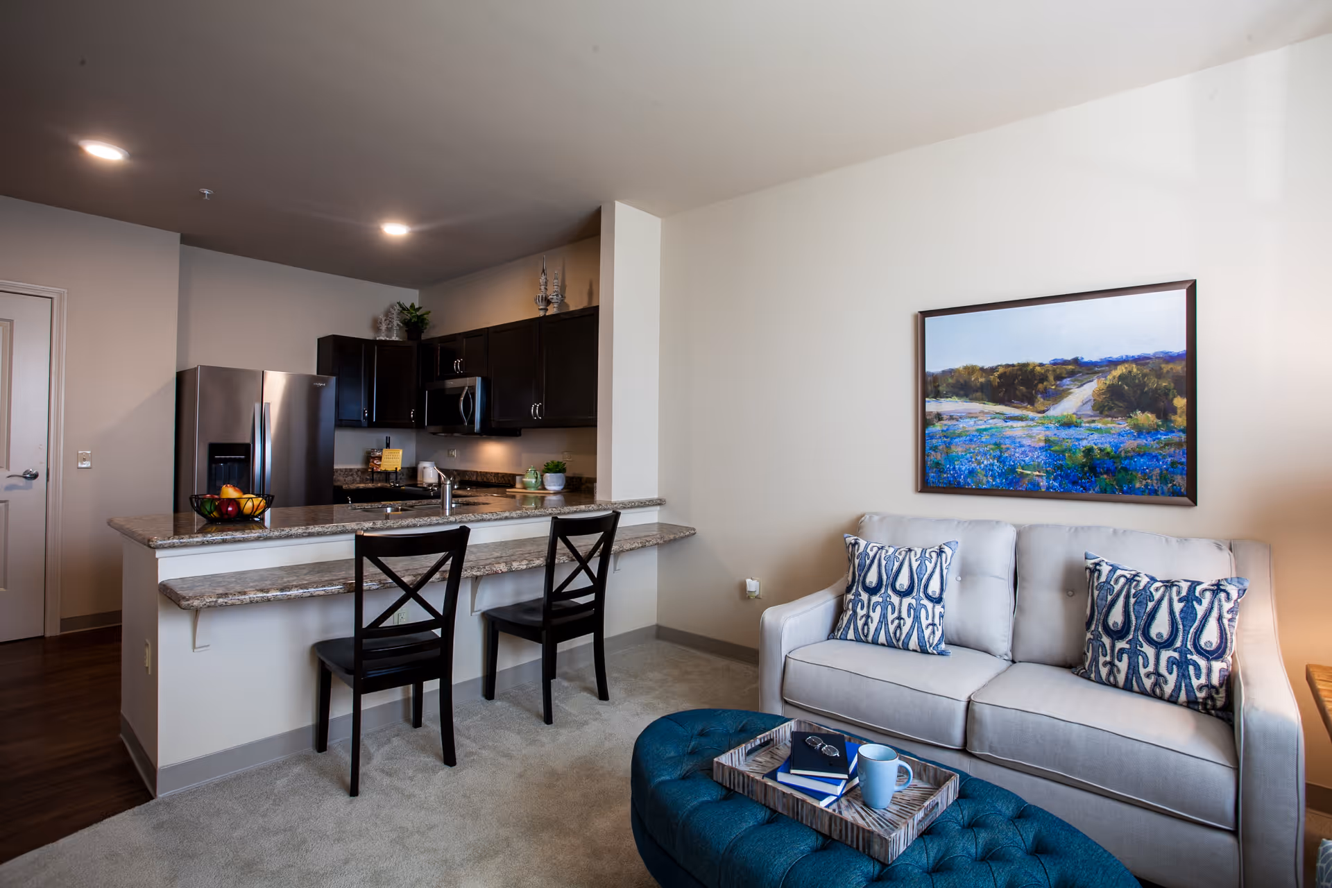 Open-plan living area with a couch and patterned pillows, a tufted ottoman coffee table, and a kitchen island with two bar stools and stainless steel appliances.