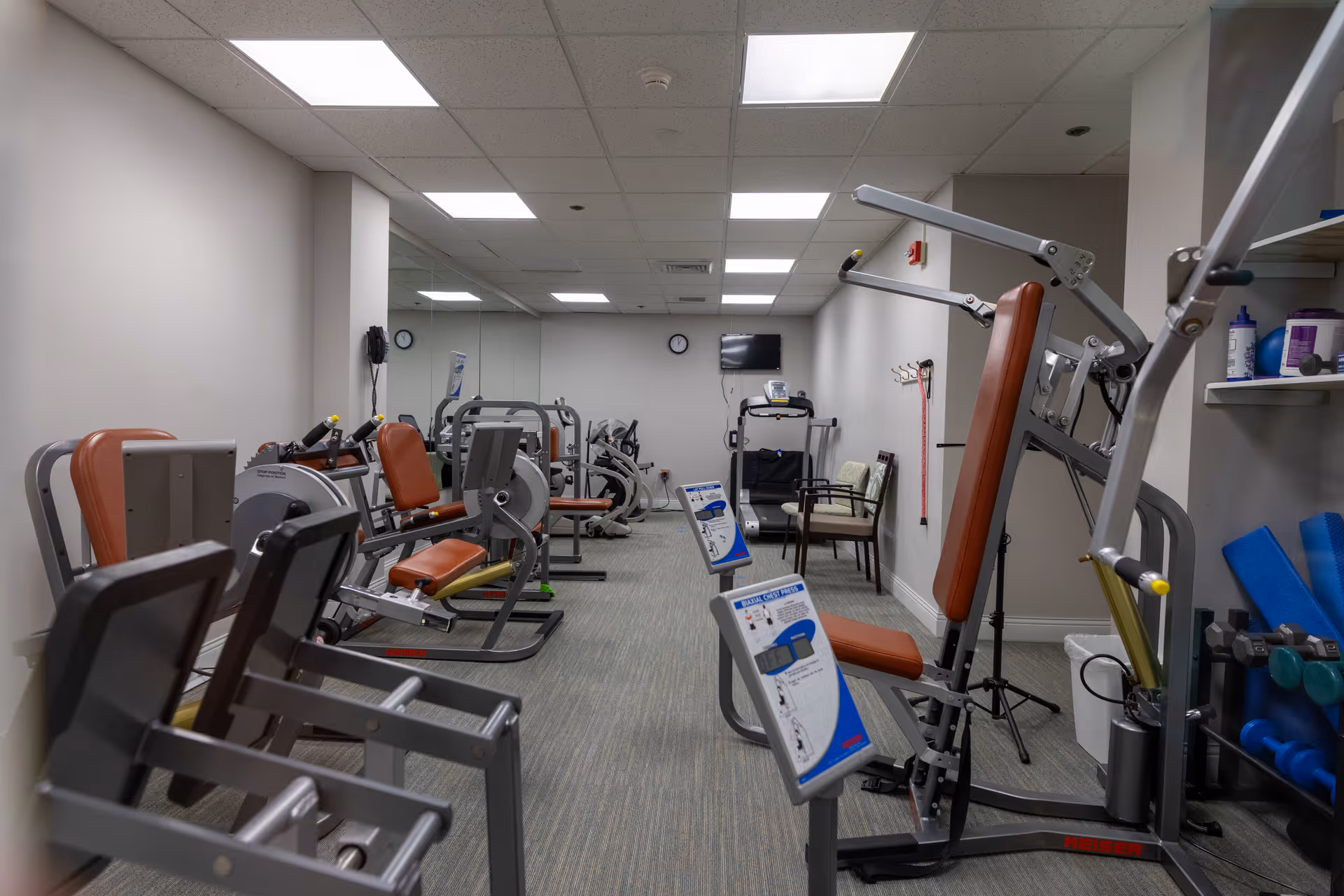 Indoor fitness room with various exercise machines including weight training equipment and cardio machines. The room has a carpeted floor, white walls, a mirrored wall at the far end, and fluorescent ceiling lights. There are chairs and a small TV mounted on the wall.