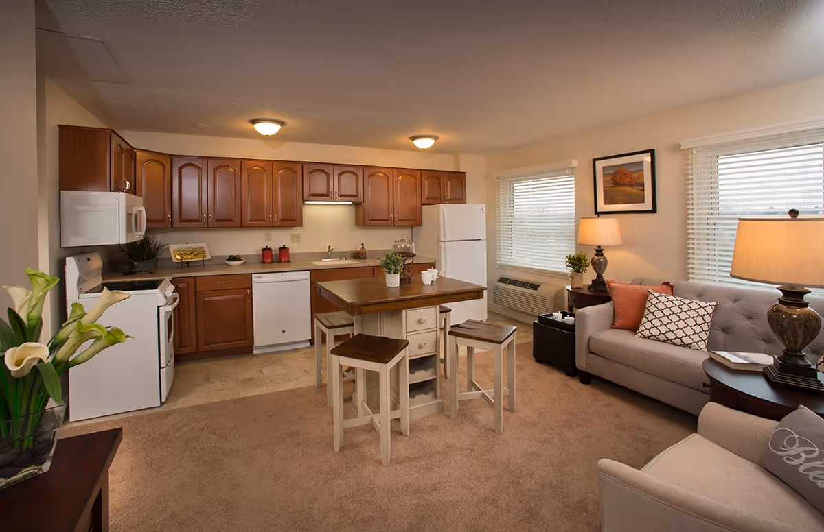A cozy living area and kitchen in an apartment at Oak Hill Village. The kitchen features wooden cabinets, a white refrigerator, stove, microwave, and dishwasher. A wooden island with four stools is in the center. The living area has a gray sofa with decorative pillows, a side table with a lamp, and a beige armchair. Two windows with blinds let in natural light, and there are framed pictures and plants adding warmth to the space.