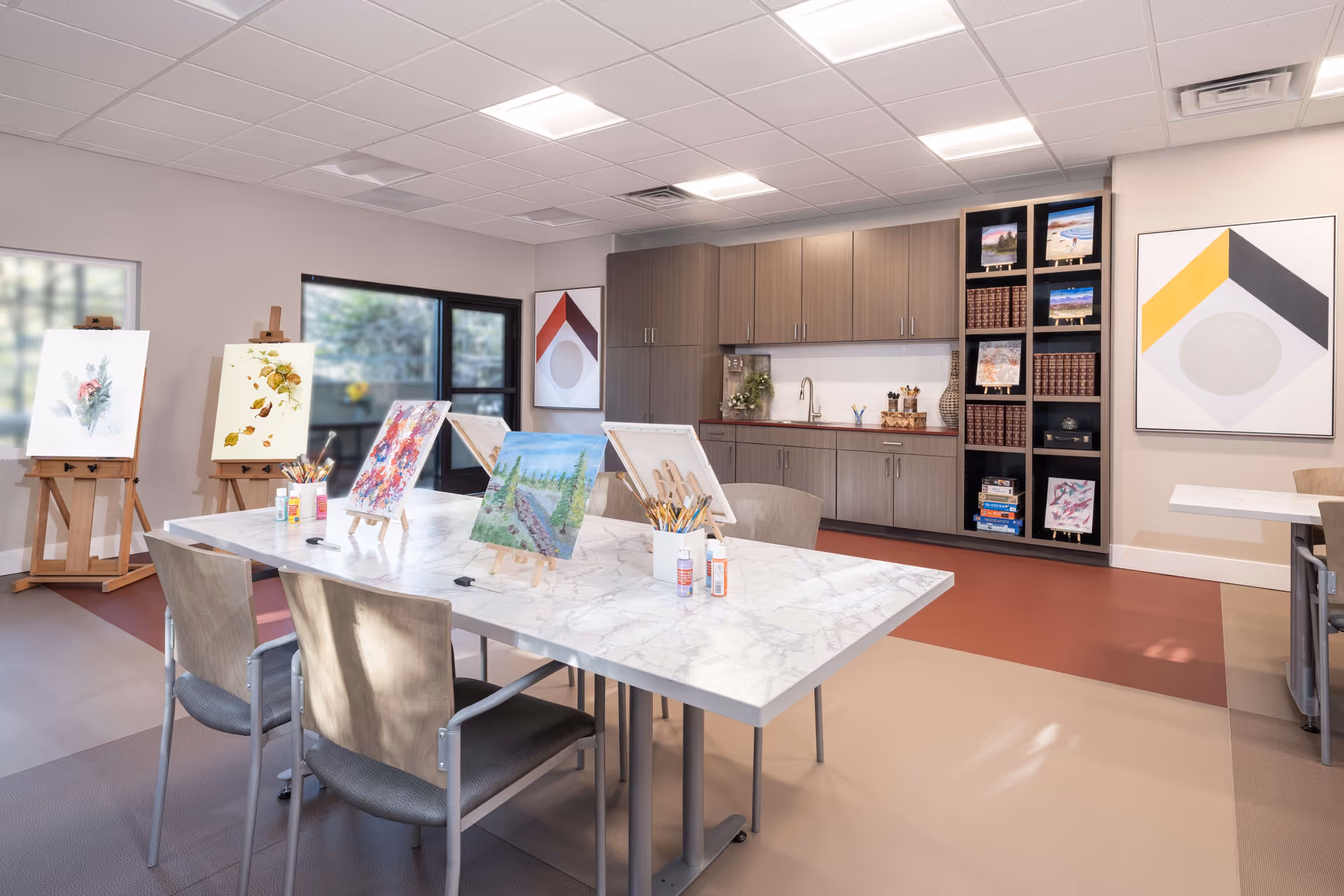 Bright art studio/activity room with tables, easels, paintings, and cabinets.